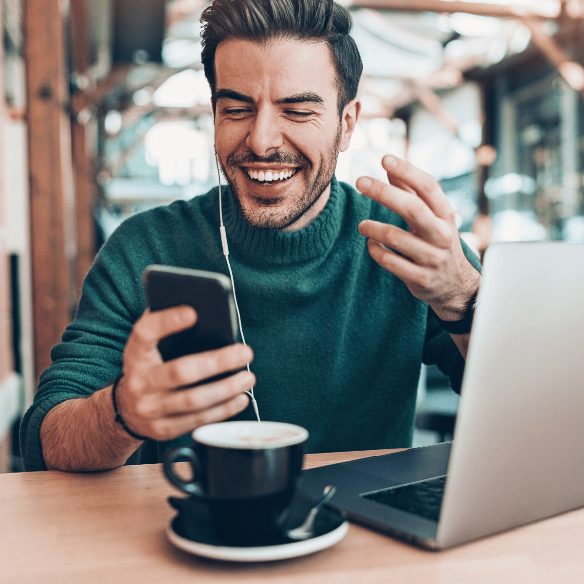 Man wearing teal sweater smiling and talking on phone with earphones in a cafe with coffee and laptop.