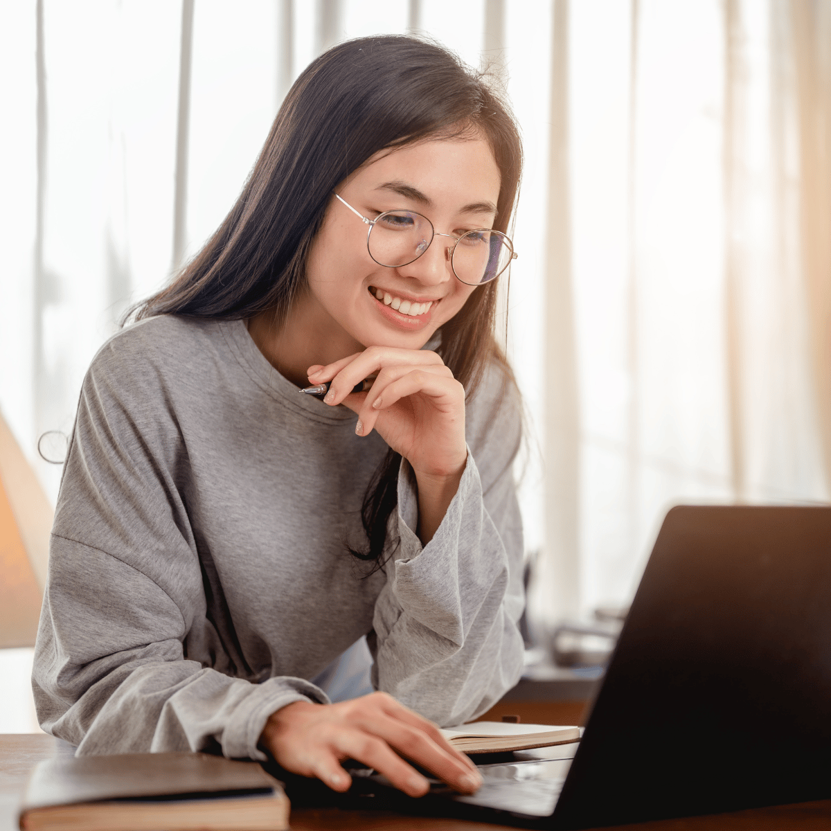 Young woman with glasses smiling while working on a laptop at a desk with books.