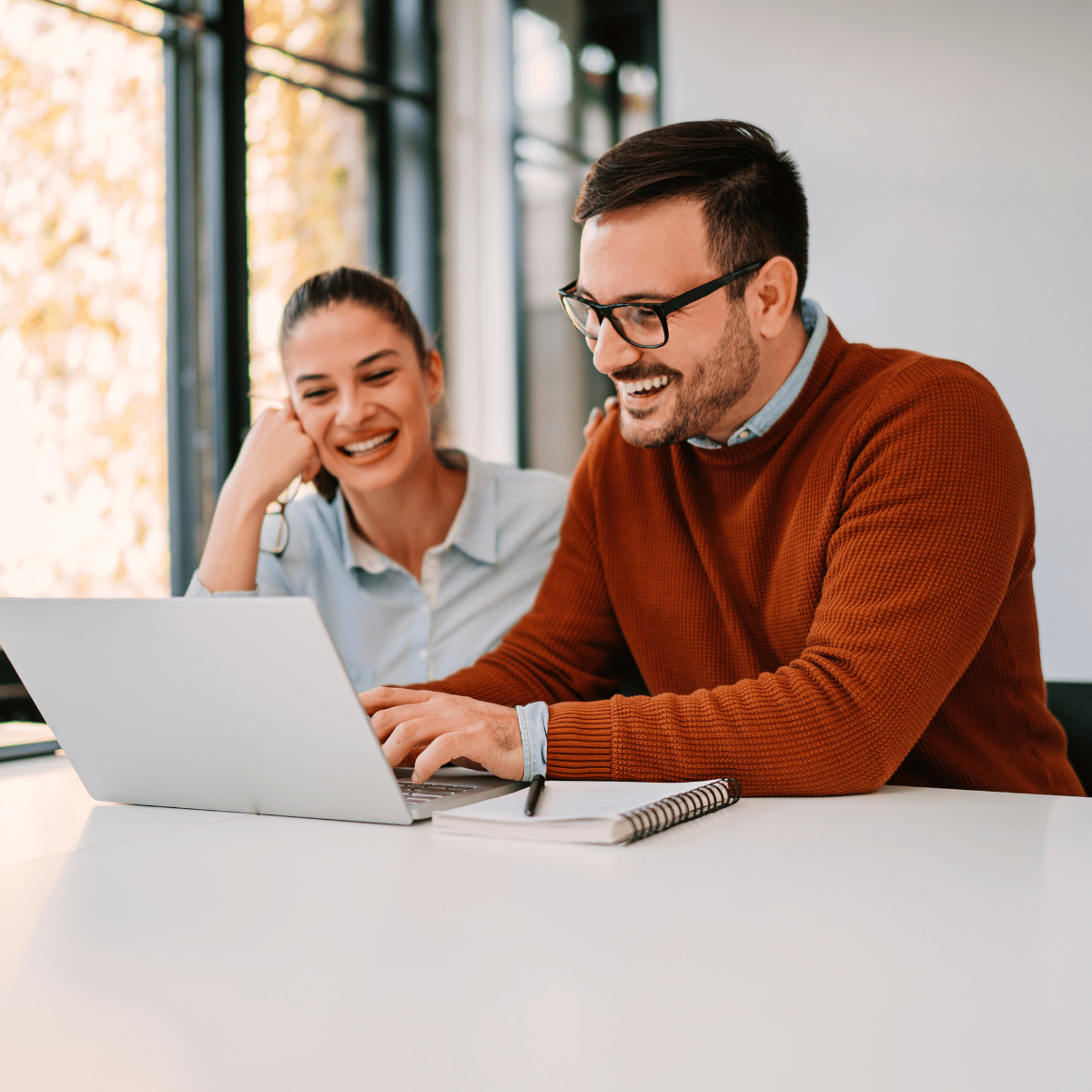 Man in orange sweater and woman in blue shirt smiling while looking at laptop screen indoors.