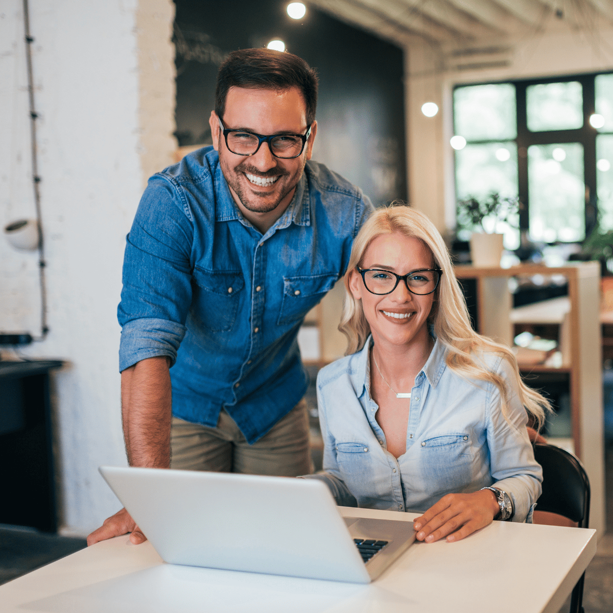 Smiling man and woman wearing glasses with laptop in modern office space.