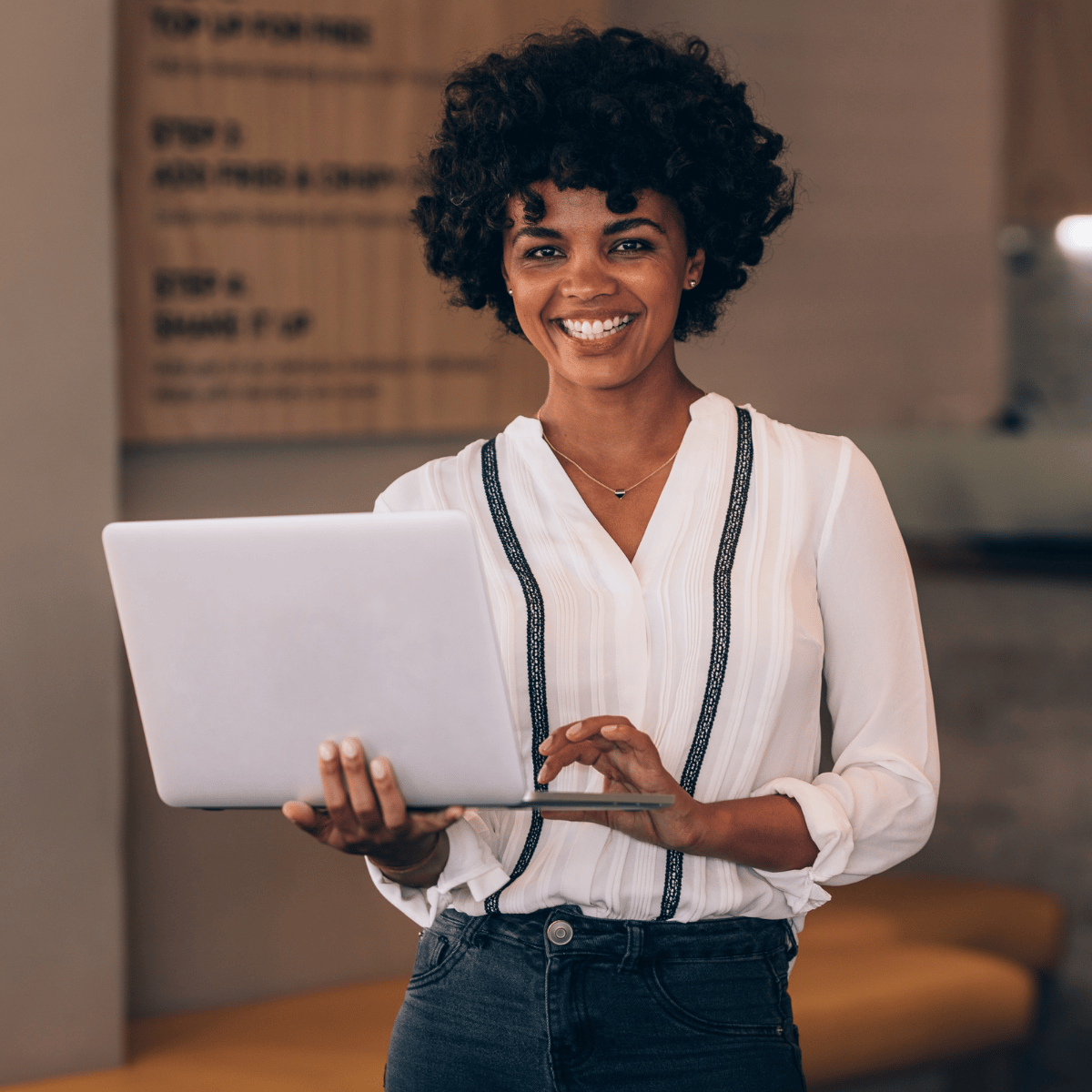Smiling woman with curly hair in white blouse holding and using a laptop indoors.