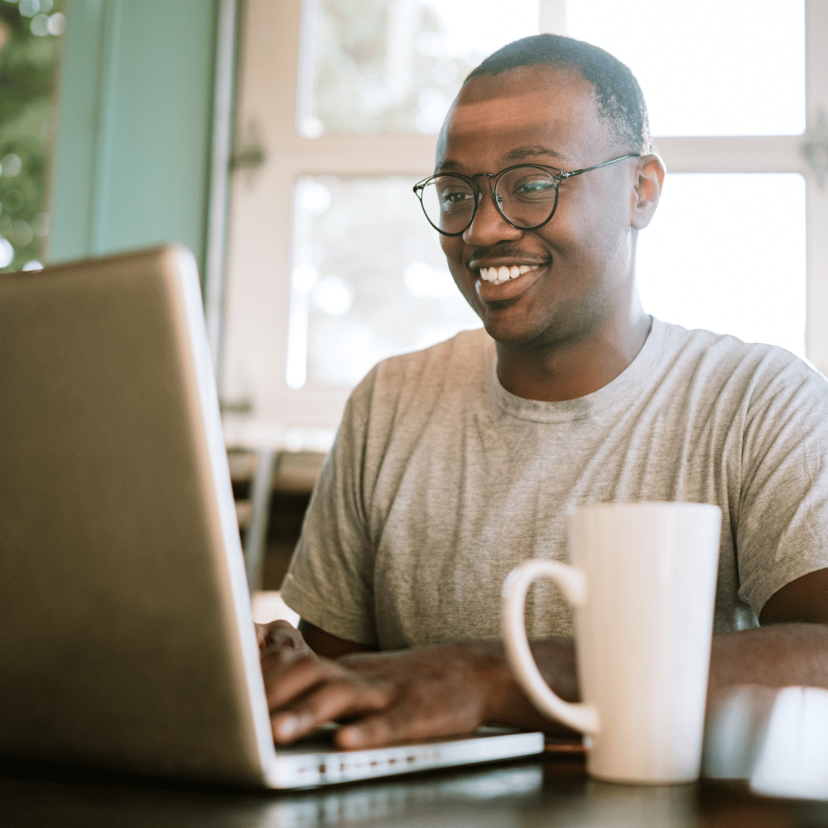 Smiling man wearing glasses and gray shirt typing on a laptop with a white mug in front.